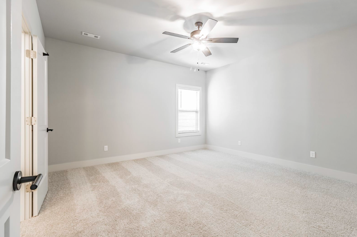 Empty bedroom with light gray walls, white trim, and beige carpet. Ceiling fan.