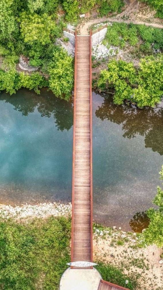 Overhead view of a narrow, brown wooden bridge spanning a turquoise river, surrounded by green trees and foliage.