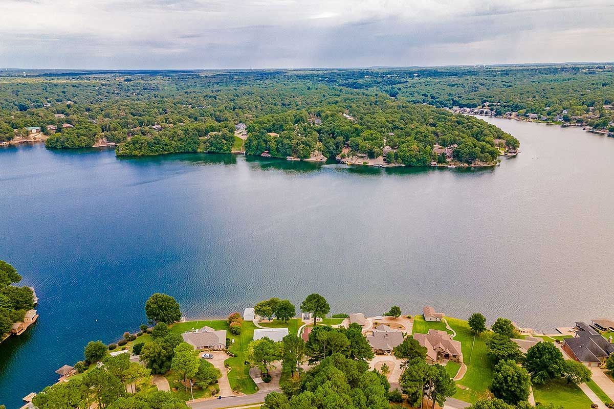 Aerial view of a lake surrounded by green trees and homes with blue water. Overcast sky.