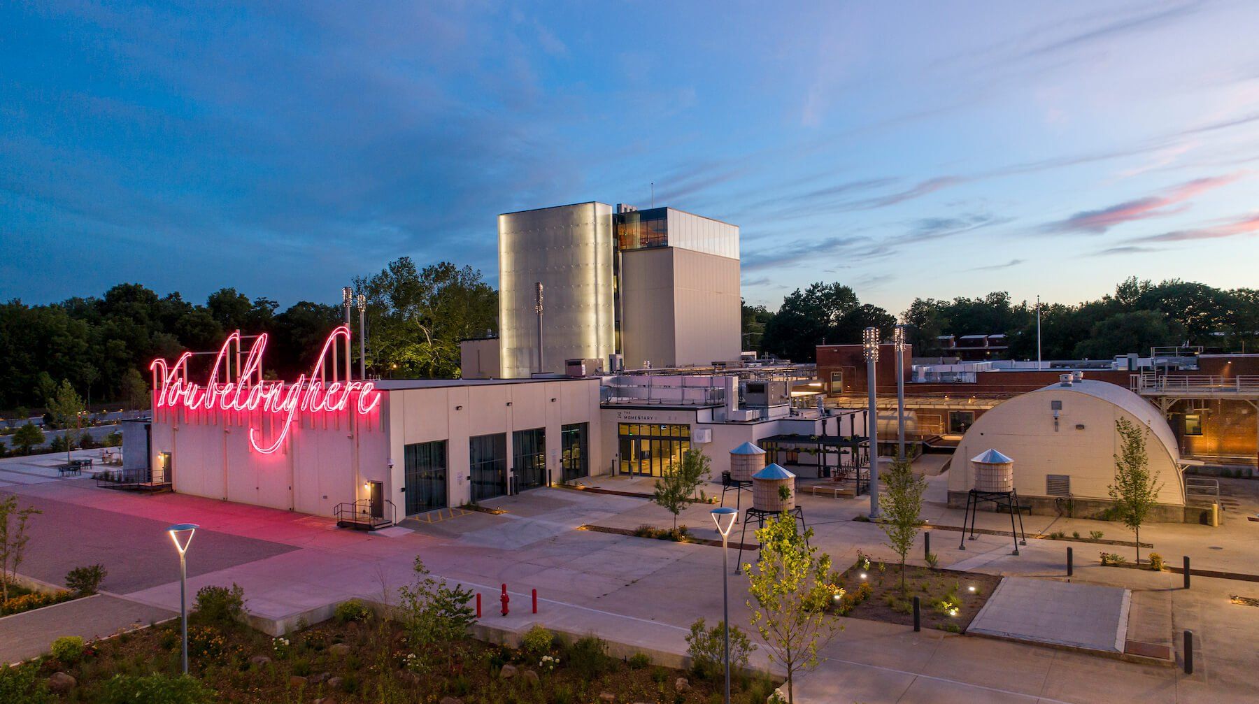 Exterior view of a modern building with a large neon sign and silos at dusk.
