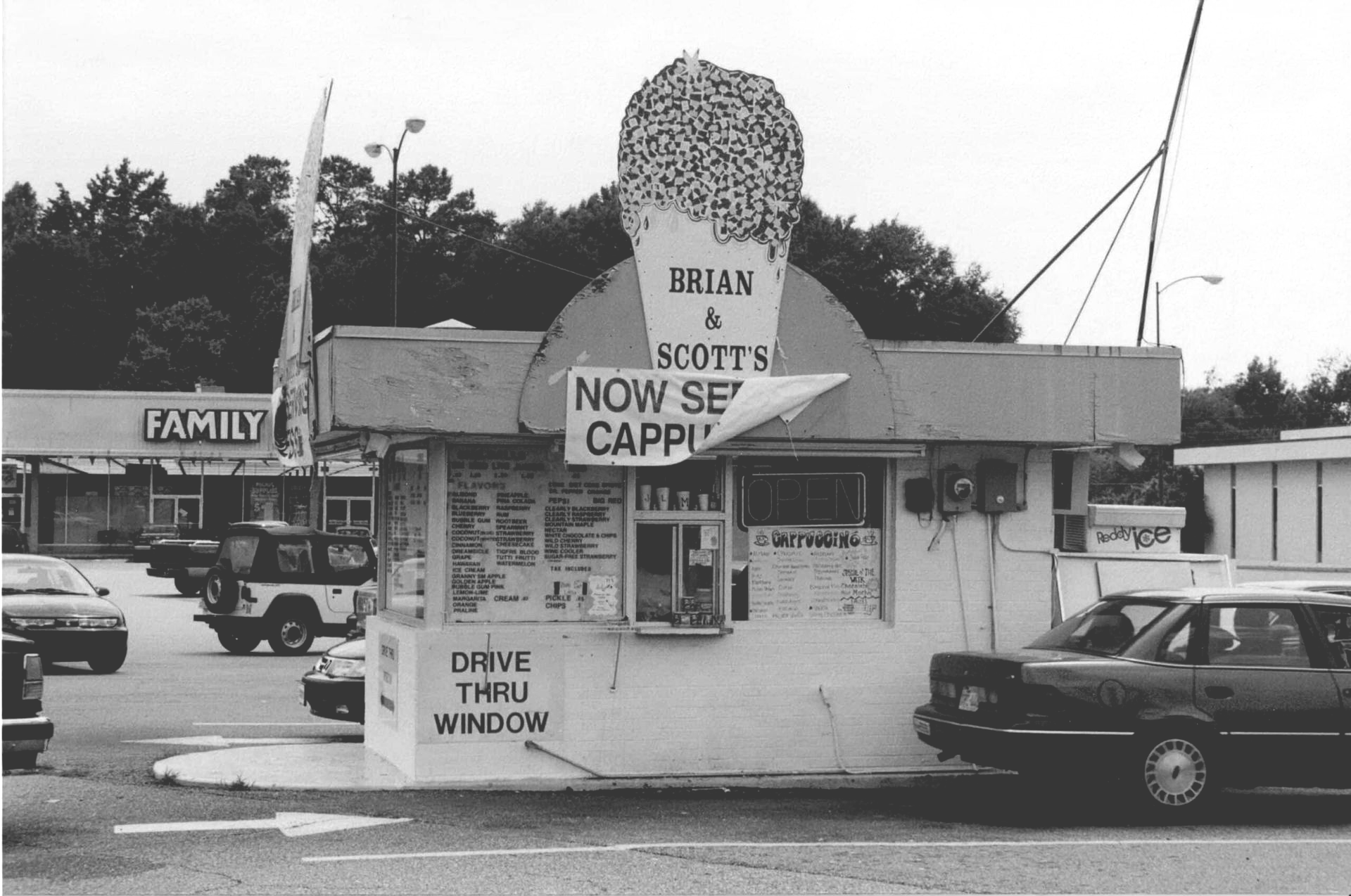 Small roadside stand with large ice cream cone sign and parked cars outside on a cloudy day
