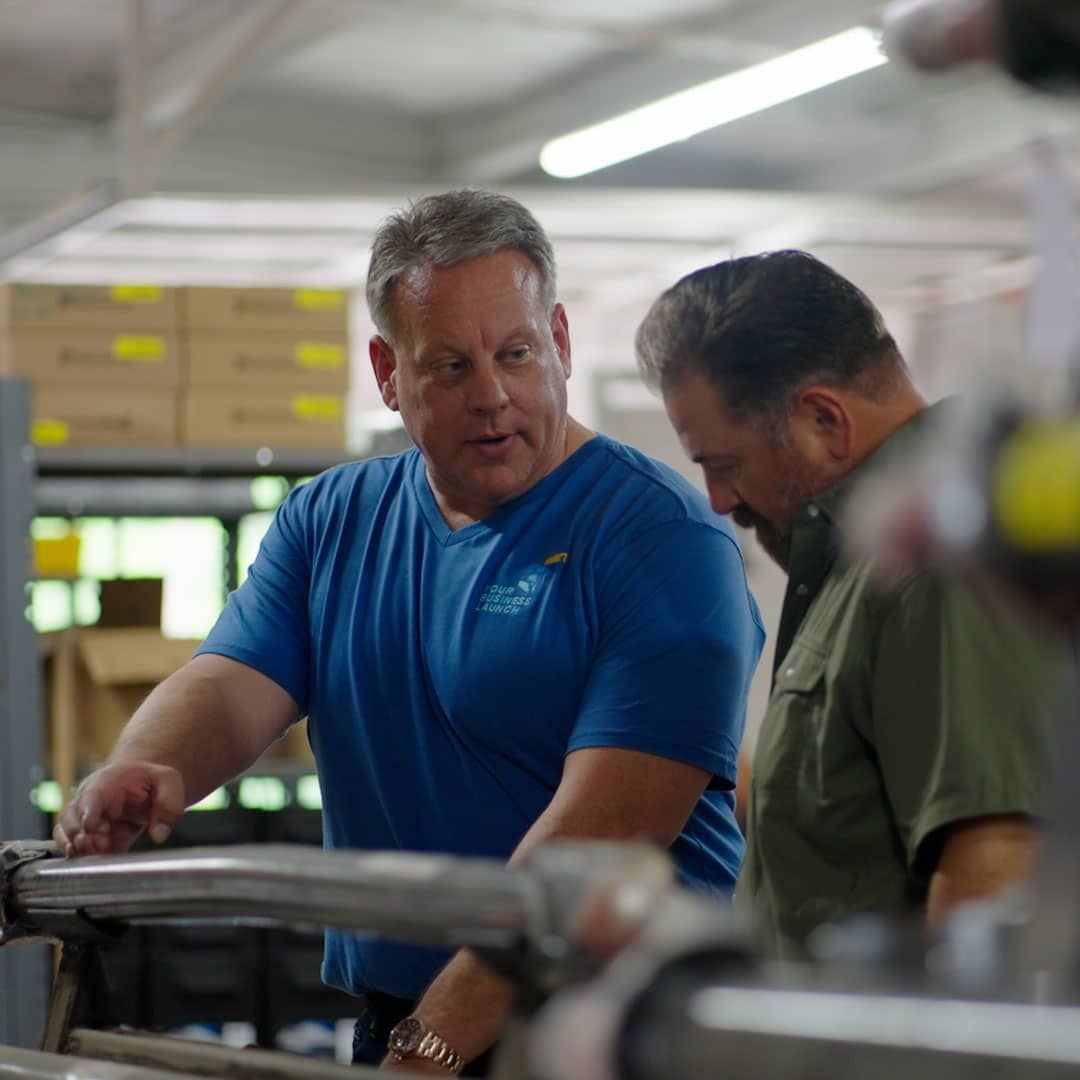 Two men in a workshop talk over a metal machine, one in a blue shirt pointing at the equipment.