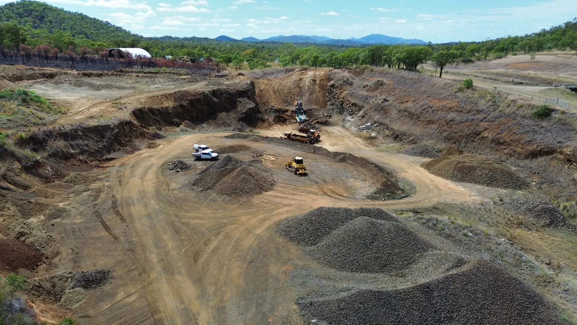 Mining operation, earthmovers, and trucks in a barren, brown landscape. Mountains and trees in the background. — Marlborough Quarry (QLD) In Marlborough, QLD