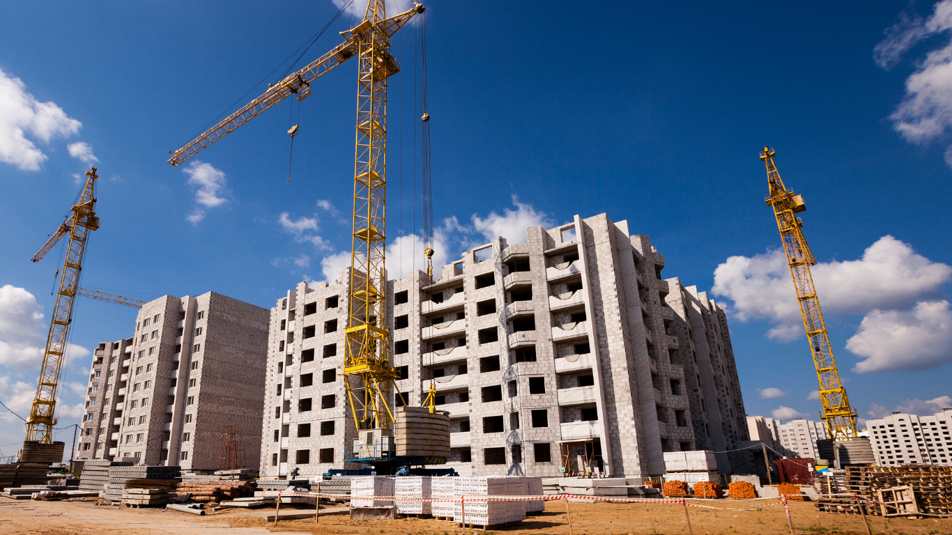 Construction site with multiple tall cranes and unfinished concrete buildings under a blue sky.