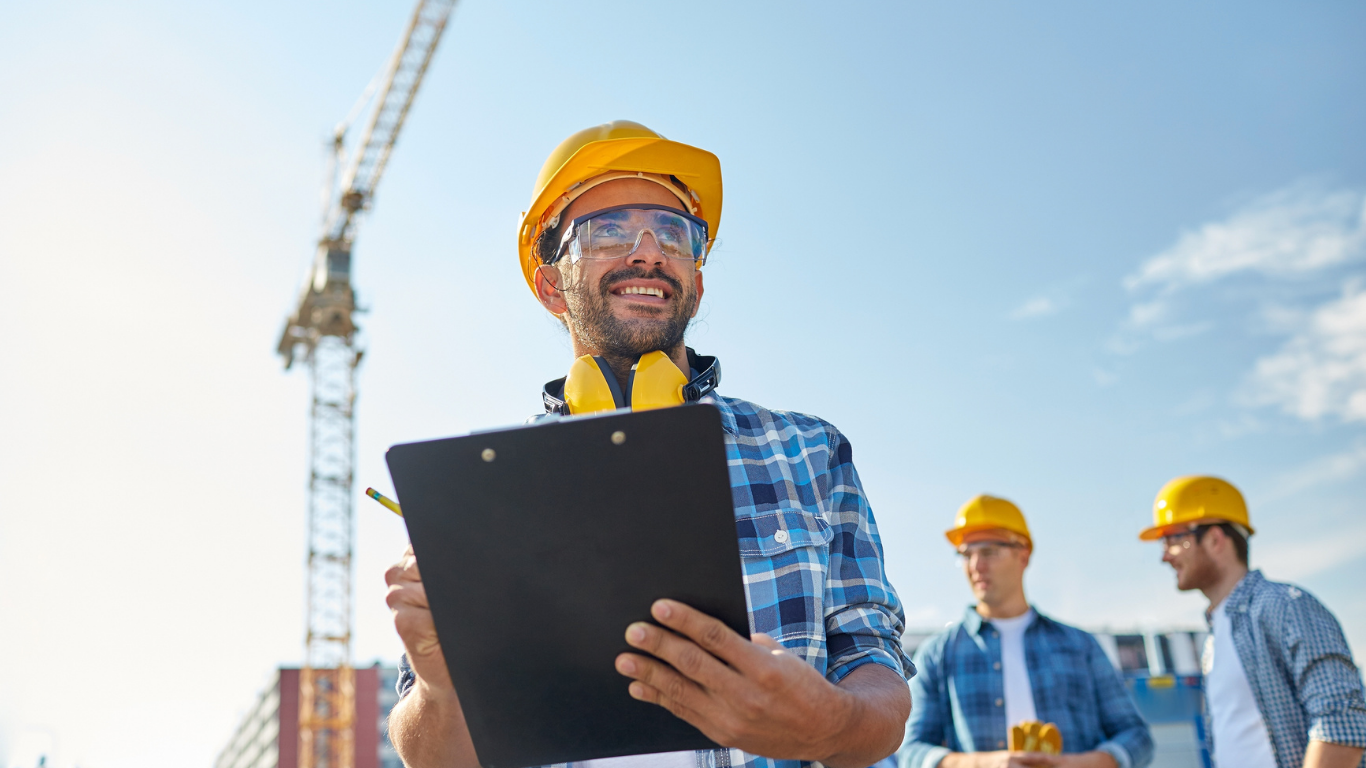 Construction worker in yellow hard hat holding a clipboard, smiling with two other workers and a crane in the background.