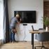 A person installs a TV on a wall. They wear a tool belt and a cap, with wires connected. A laptop sits on a nearby table.