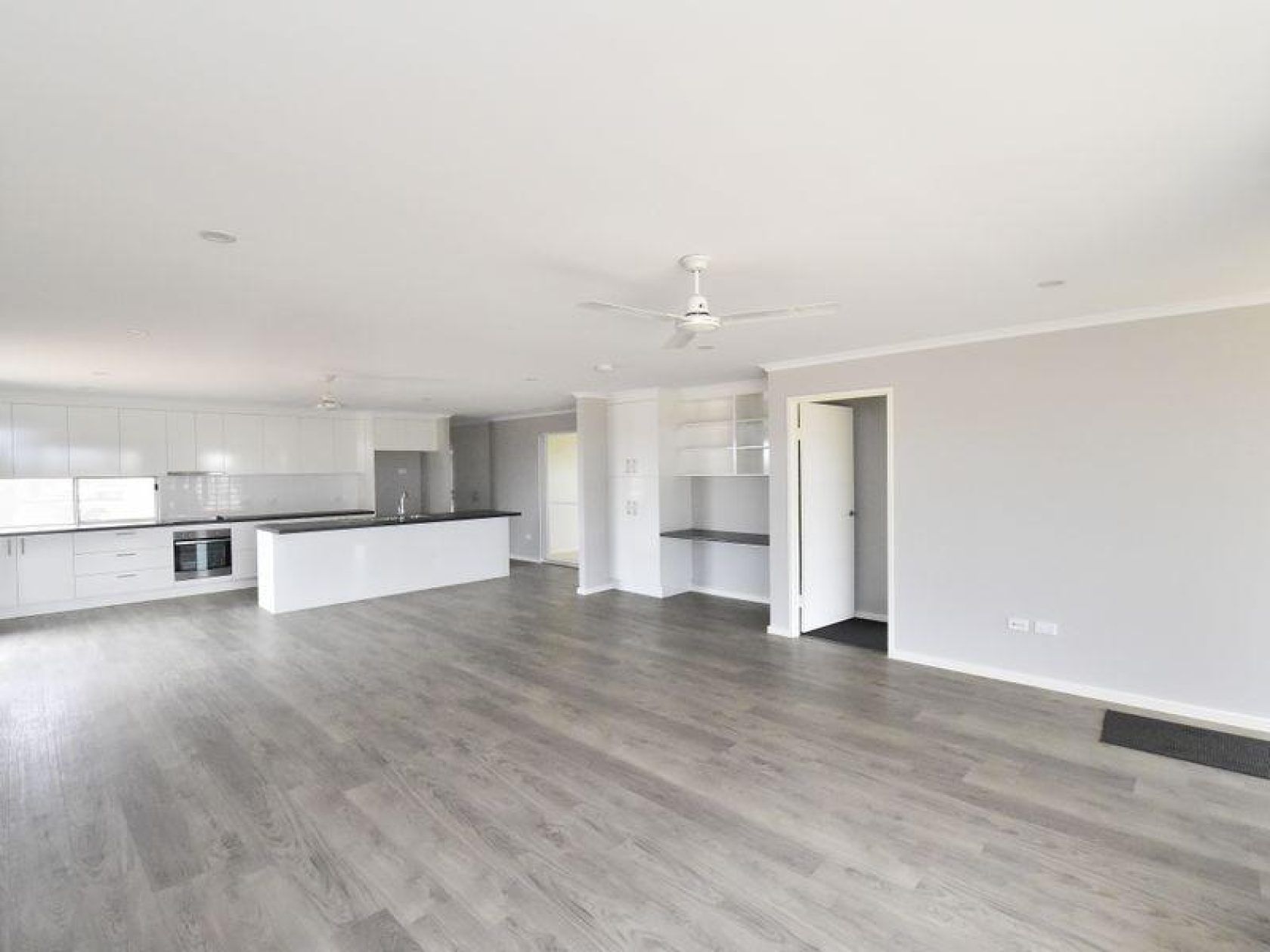 Open-plan living space with kitchen and doorway. Light grey walls, flooring, and white cabinets.