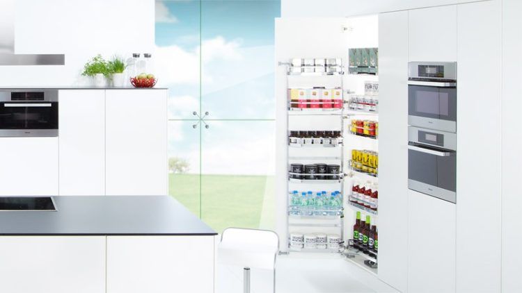 Modern white kitchen with a pull-out pantry revealing food items, next to ovens and a stovetop.