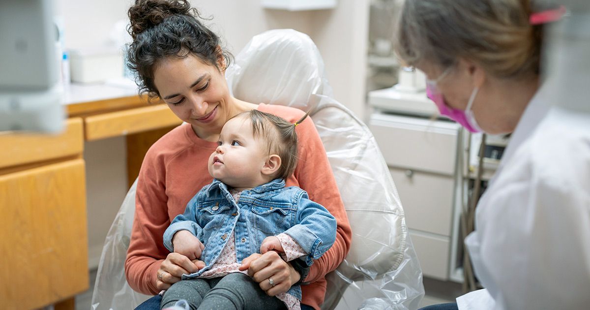 A woman is holding a baby in a dental chair.