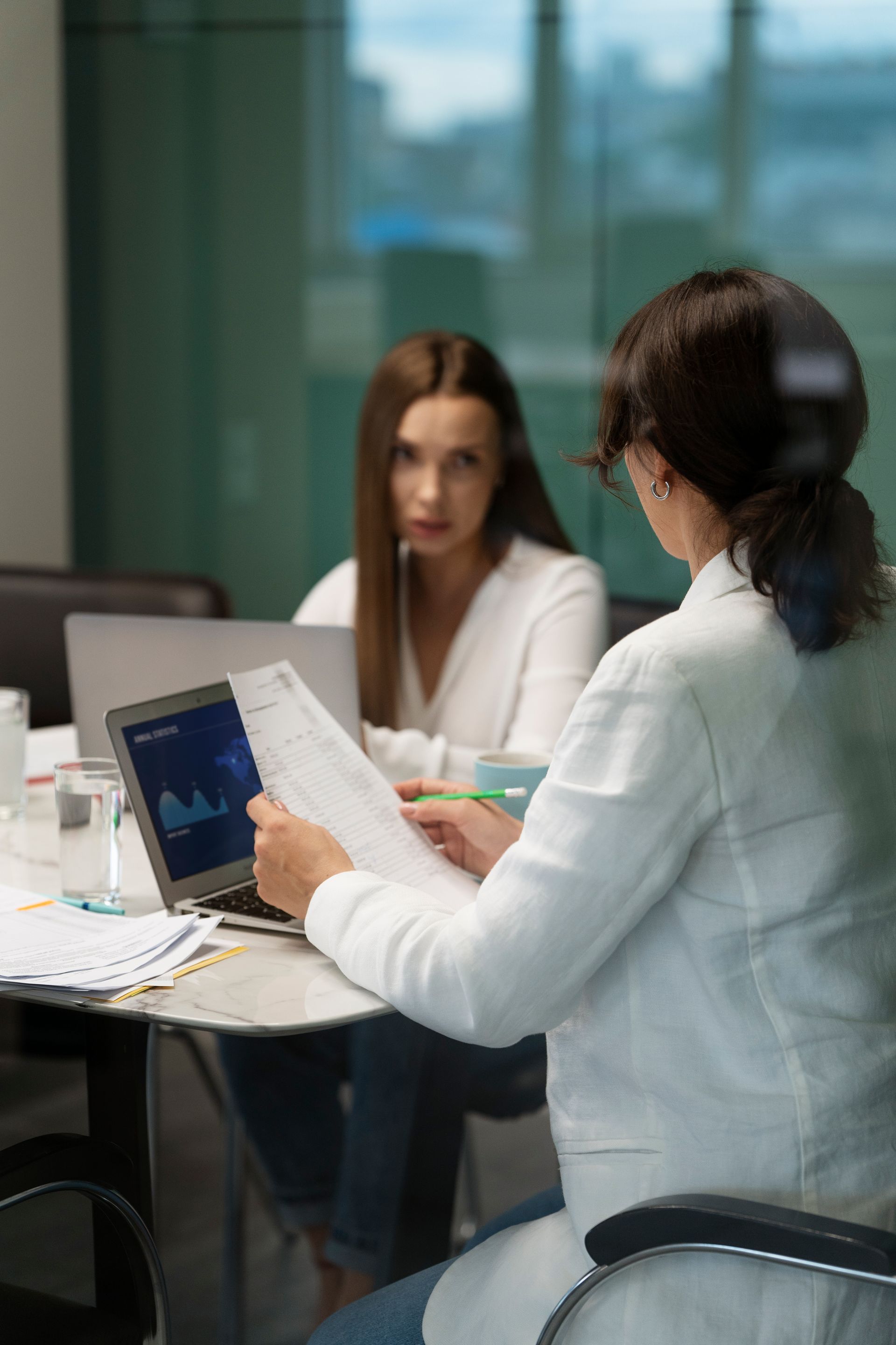 Two women are sitting at a table with laptops and papers.