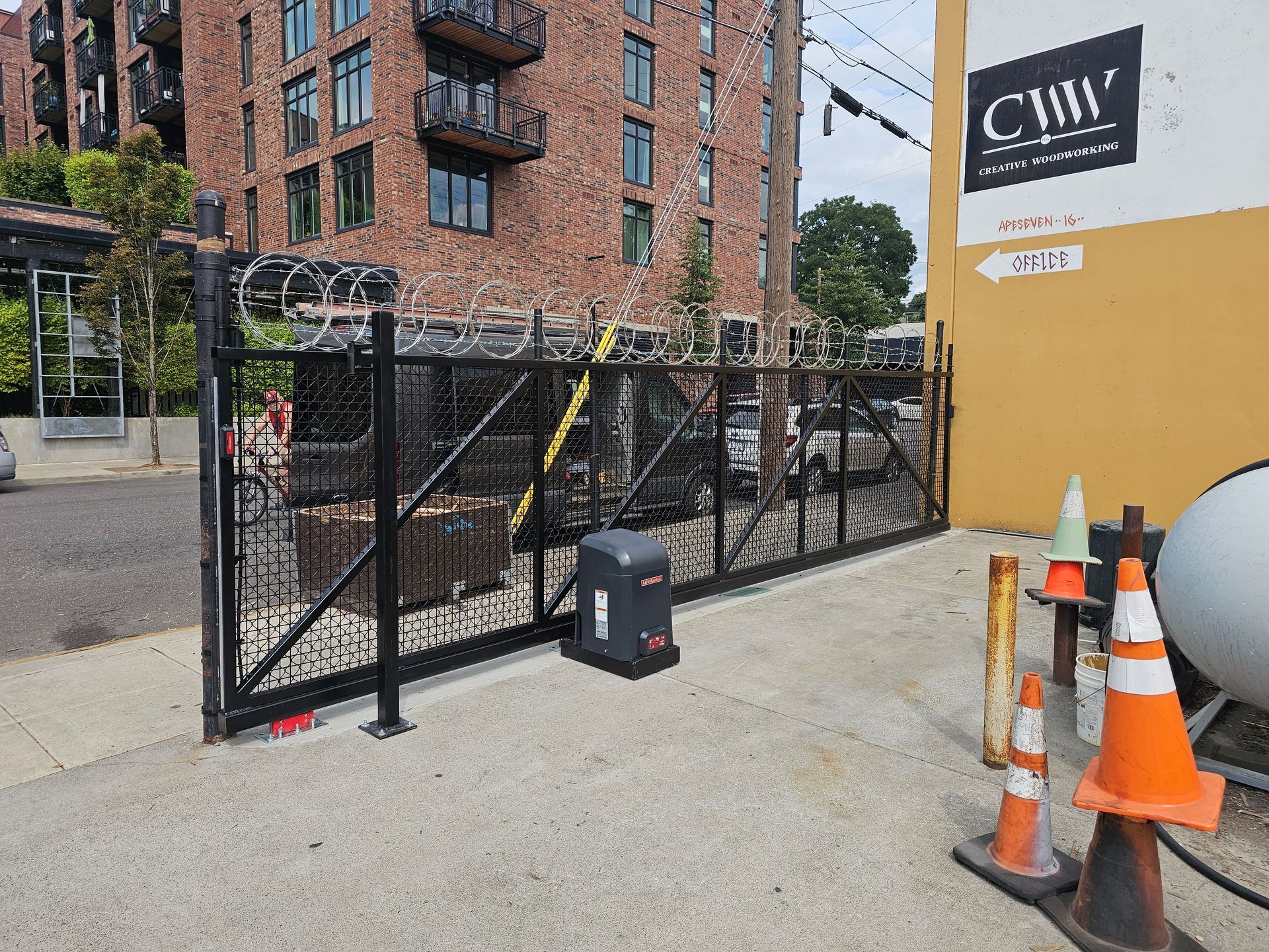 Black gate with razor wire, automatic opener, brick building background. Orange cones and sign.