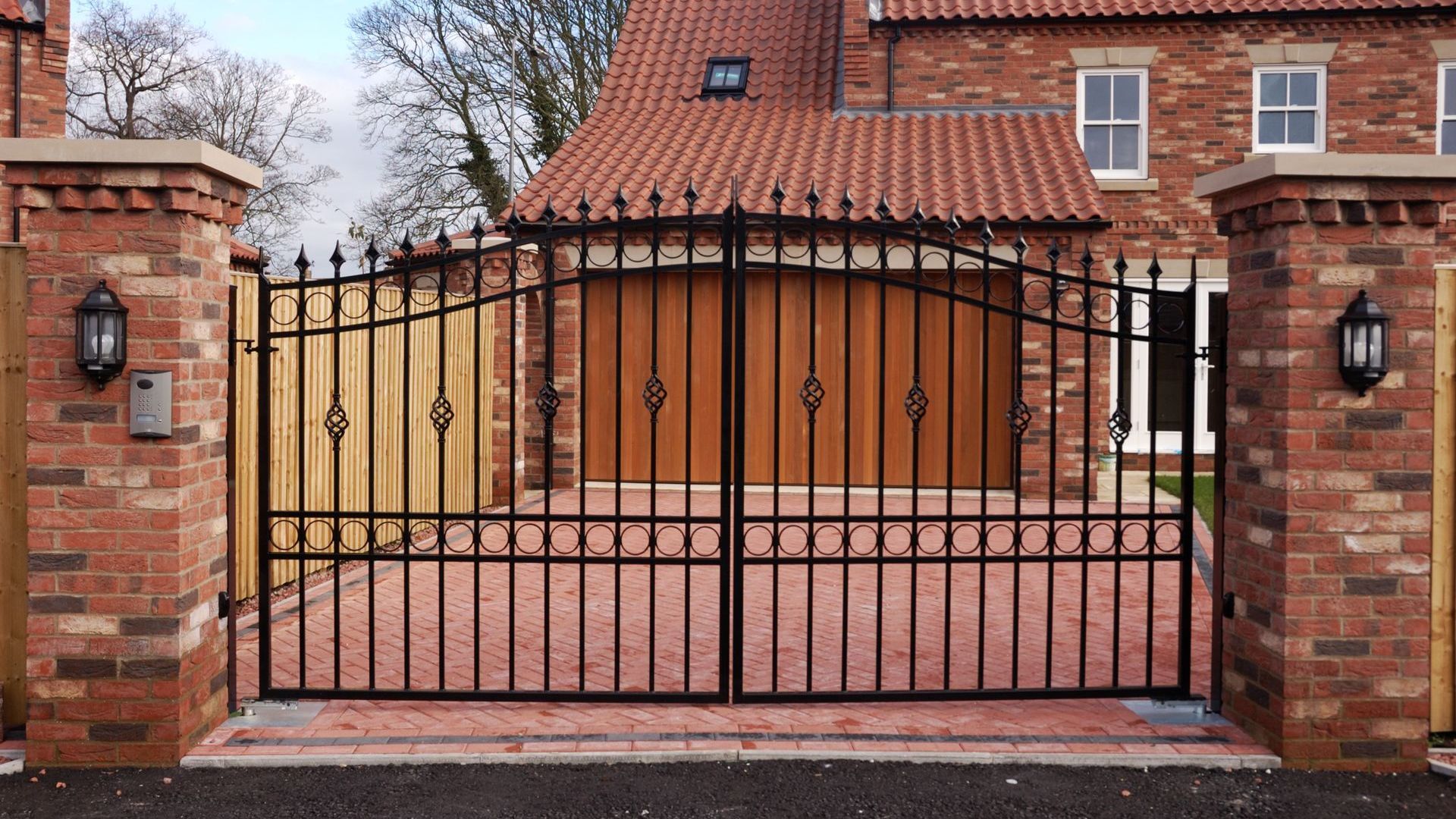 Black wrought-iron driveway gates between brick pillars, leading to a house with a red tile roof.