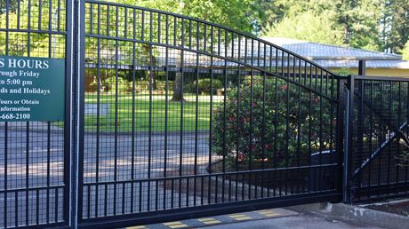 Black metal gate with arched top, leading into a green space with a sign indicating hours. Black metal gate with arched top, leading into a green space with a sign indicating hours.