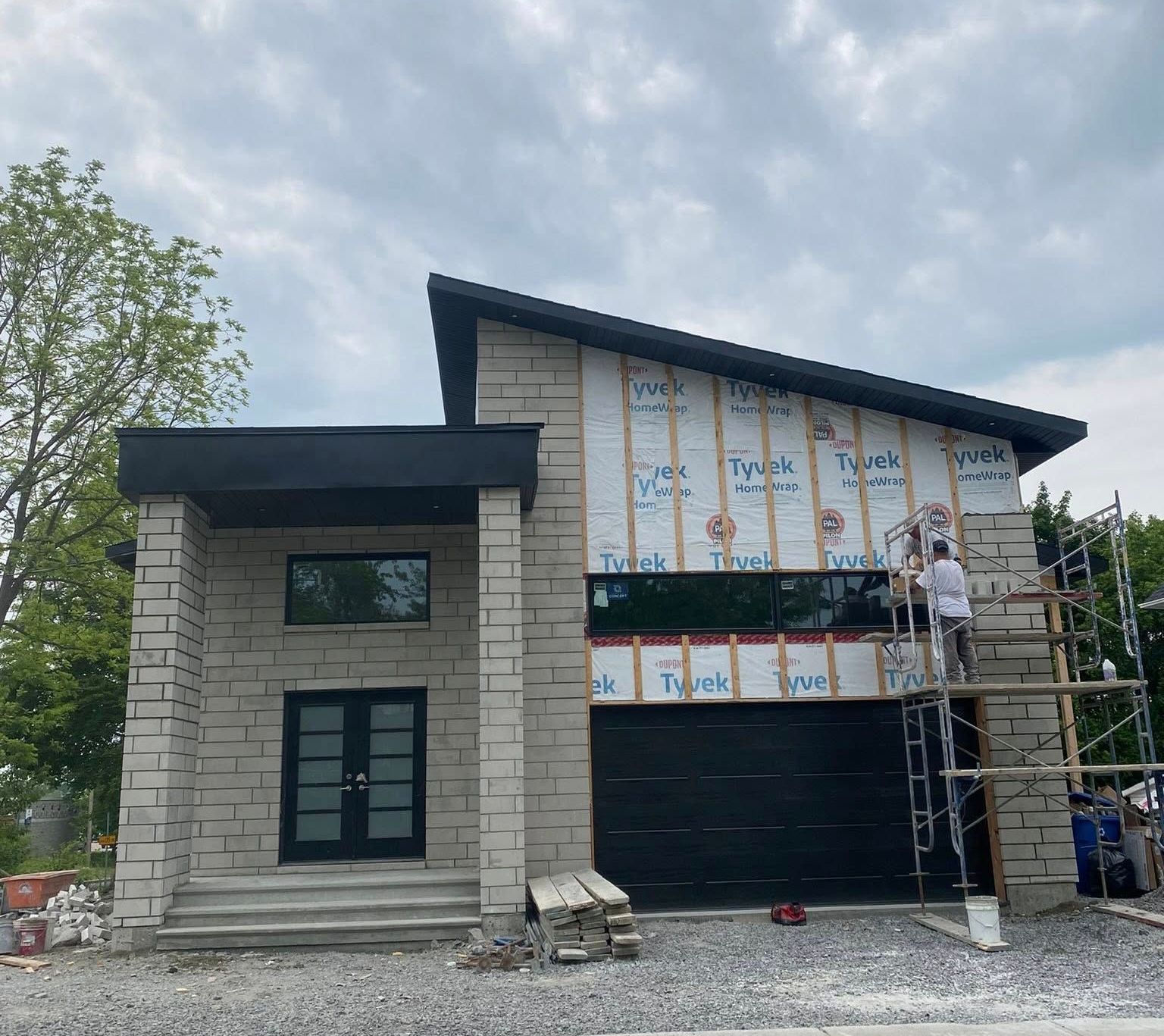 Maison moderne en construction avec façade en pierre, porte de garage noire et ouvriers sur échafaudage.