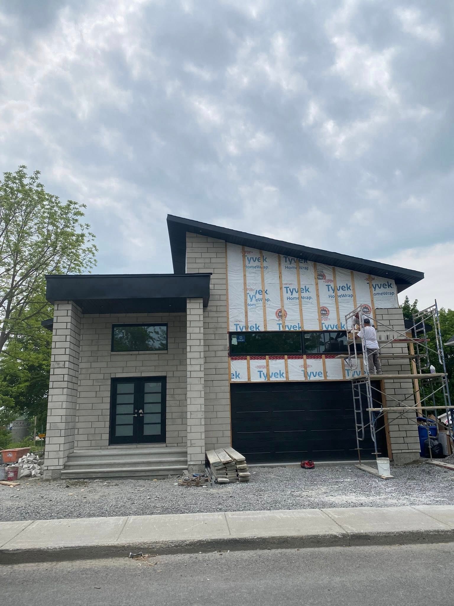 Maison moderne en construction avec ouvriers sur échafaudage. Extérieur avec porte de garage noire et façade en pierre grise.