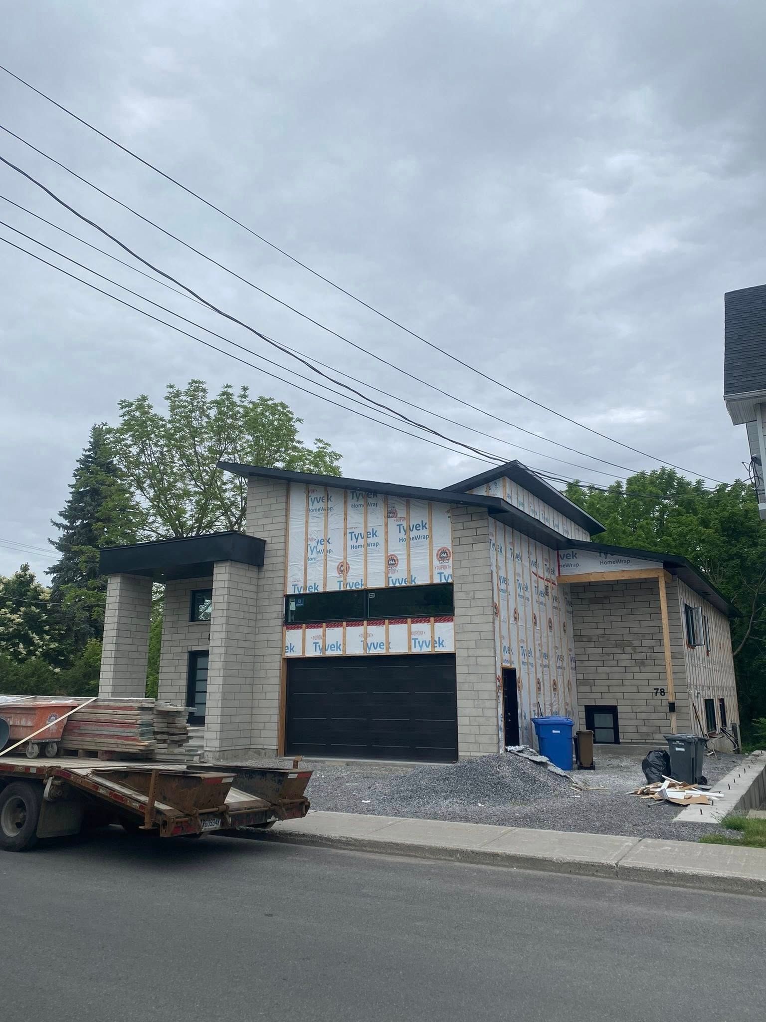 Maison en construction; extérieur en briques grises, porte de garage noire, poubelles bleues sur allée en gravier.