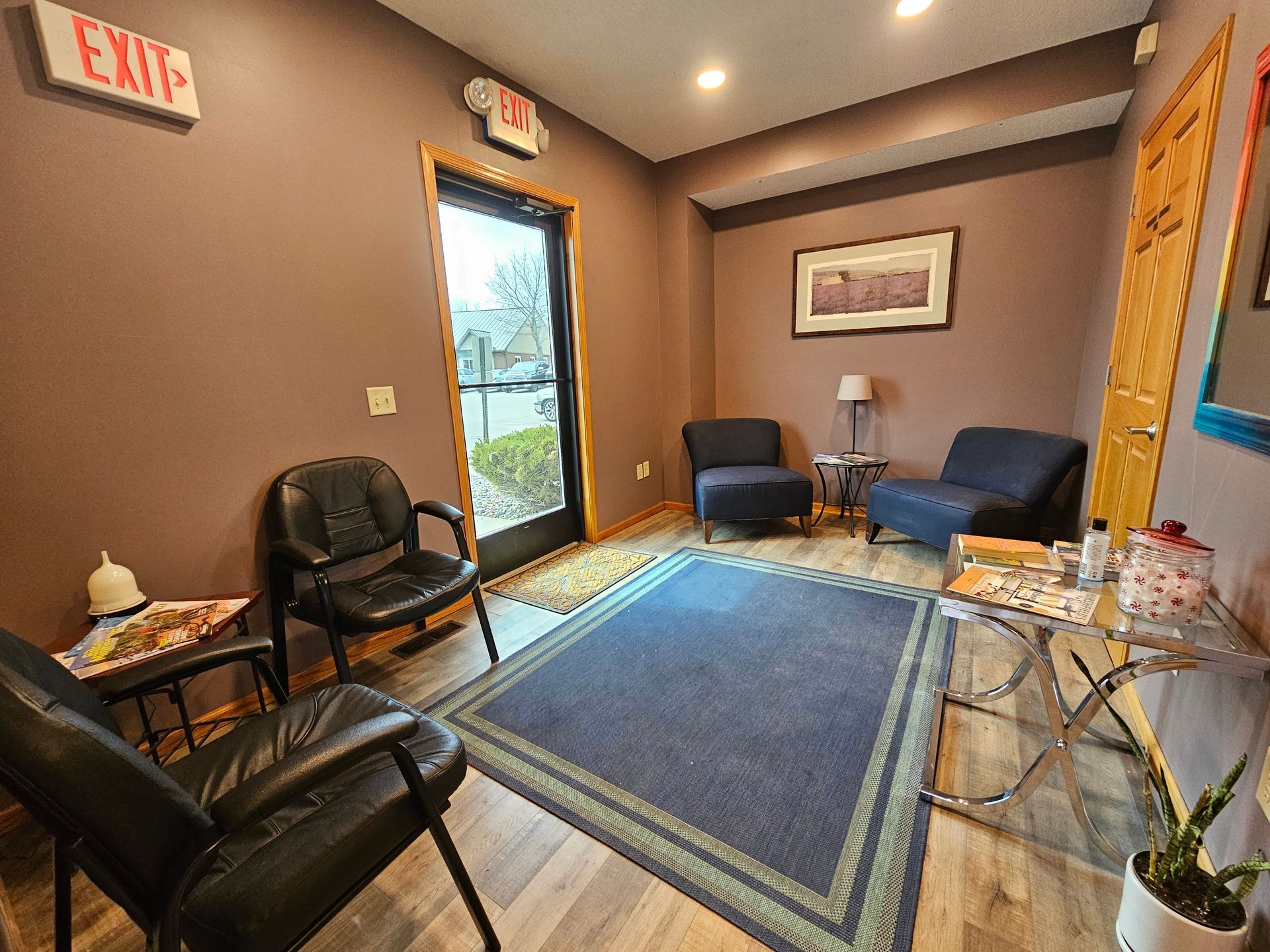 Waiting room with blue rug, two black chairs, and two blue armchairs. Brown walls and exit signs.