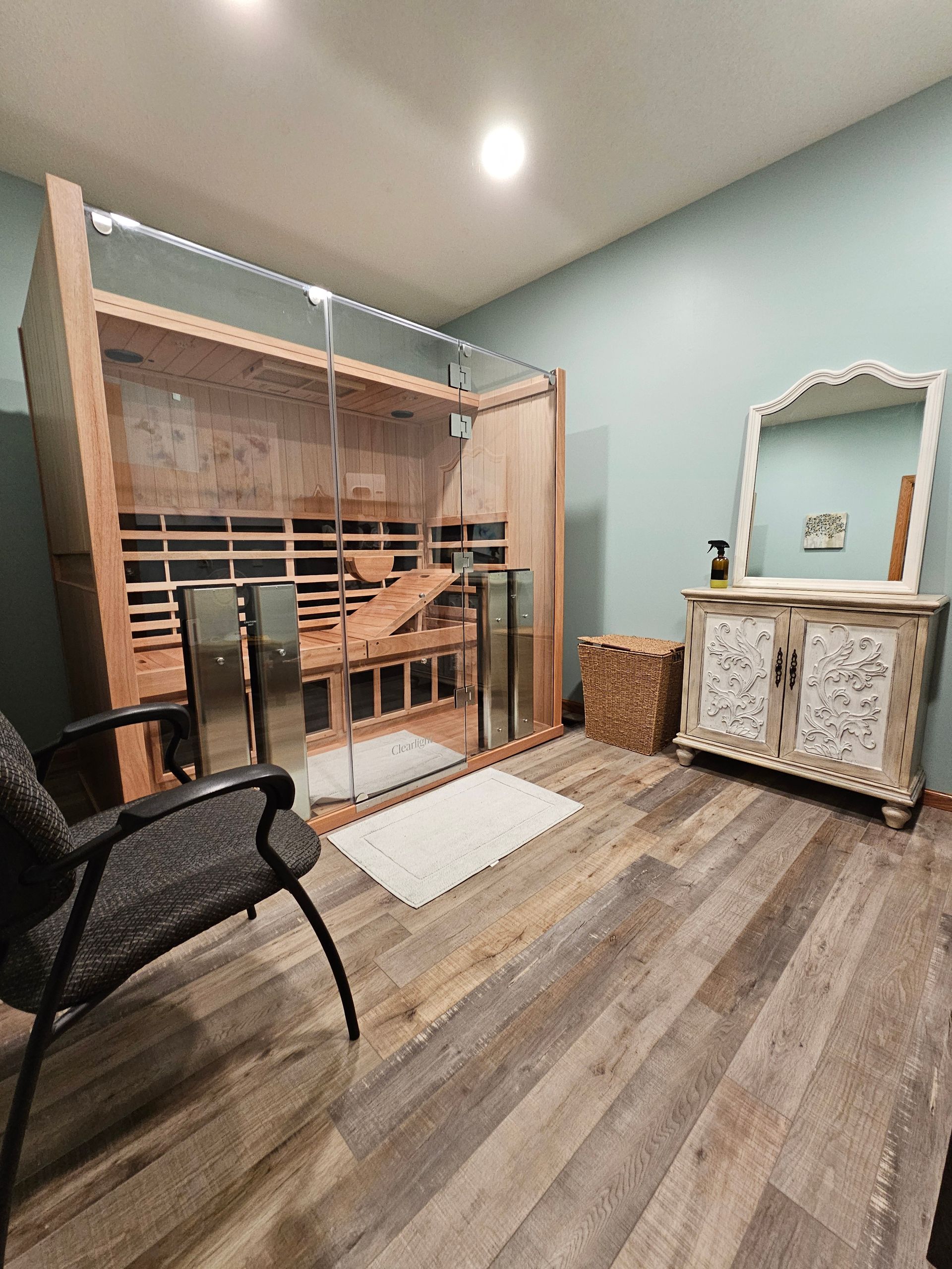 A sauna with a wood interior and glass door, in a room with light blue walls, a chair, and a decorative cabinet.