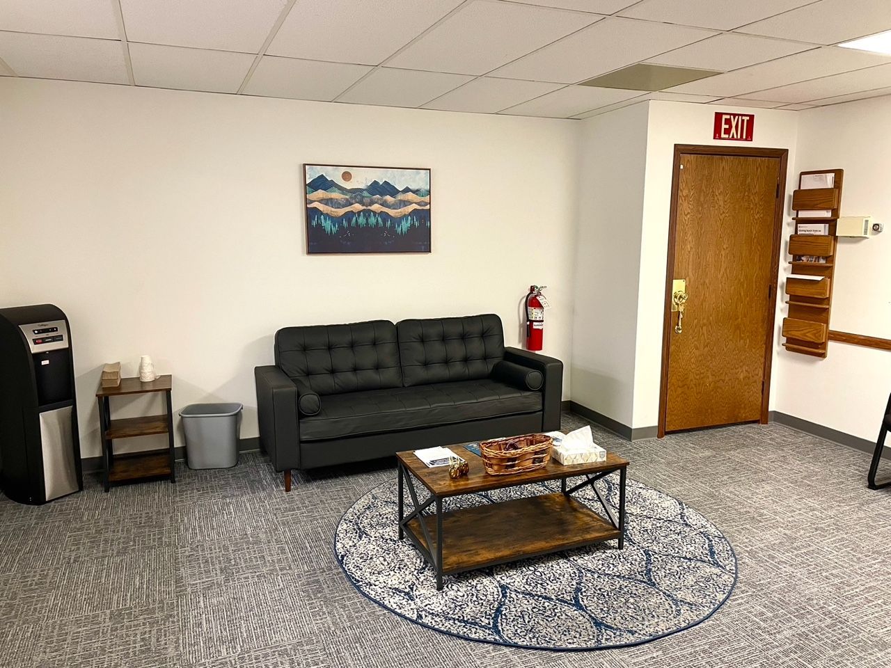 Waiting room with gray sofa, coffee table, water cooler, art, and exit door.