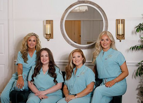 Four people in teal scrubs posing for a photo. A large mirror and sconces are visible on the wall.