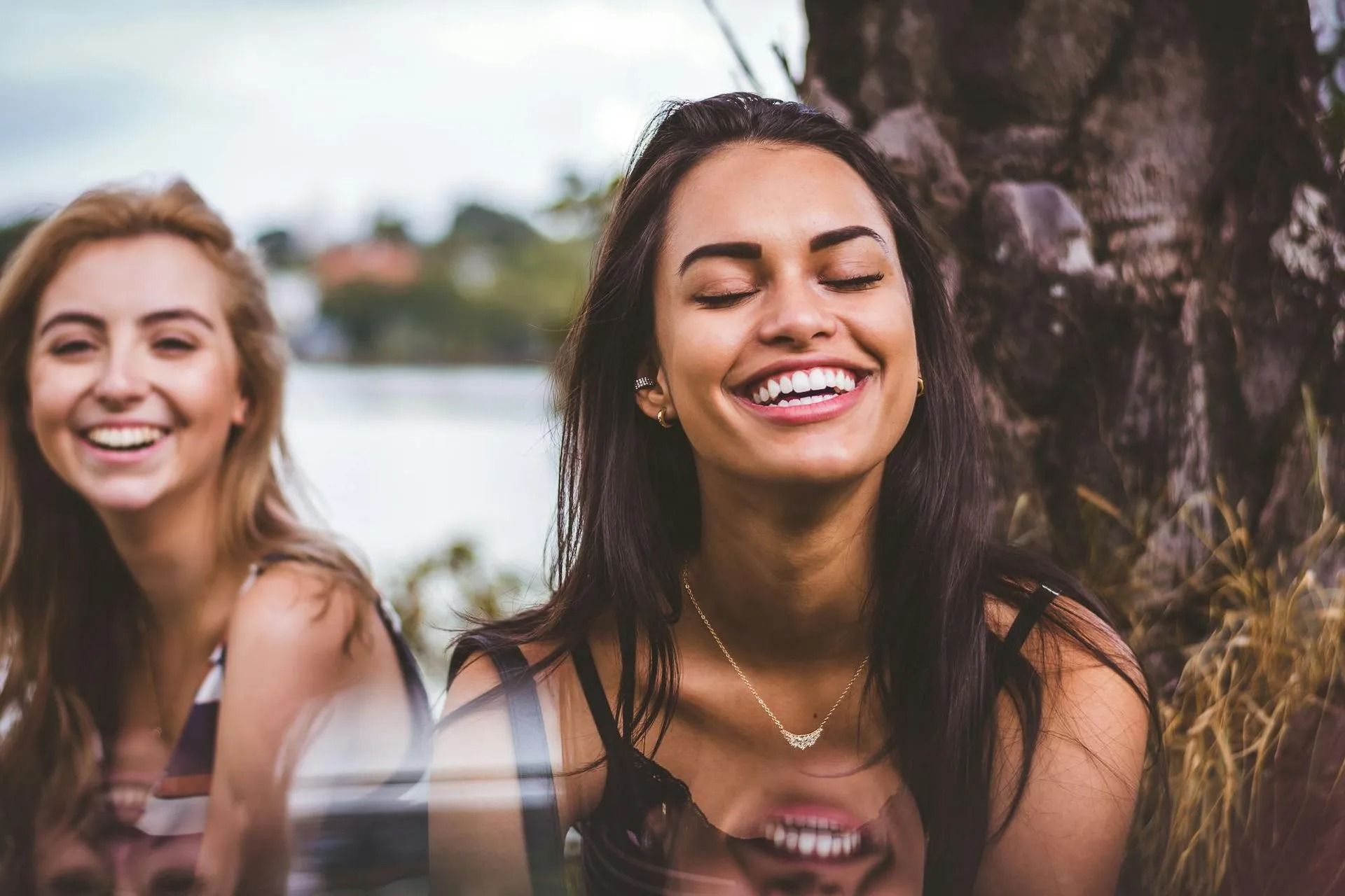Two smiling women outdoors near water and trees.