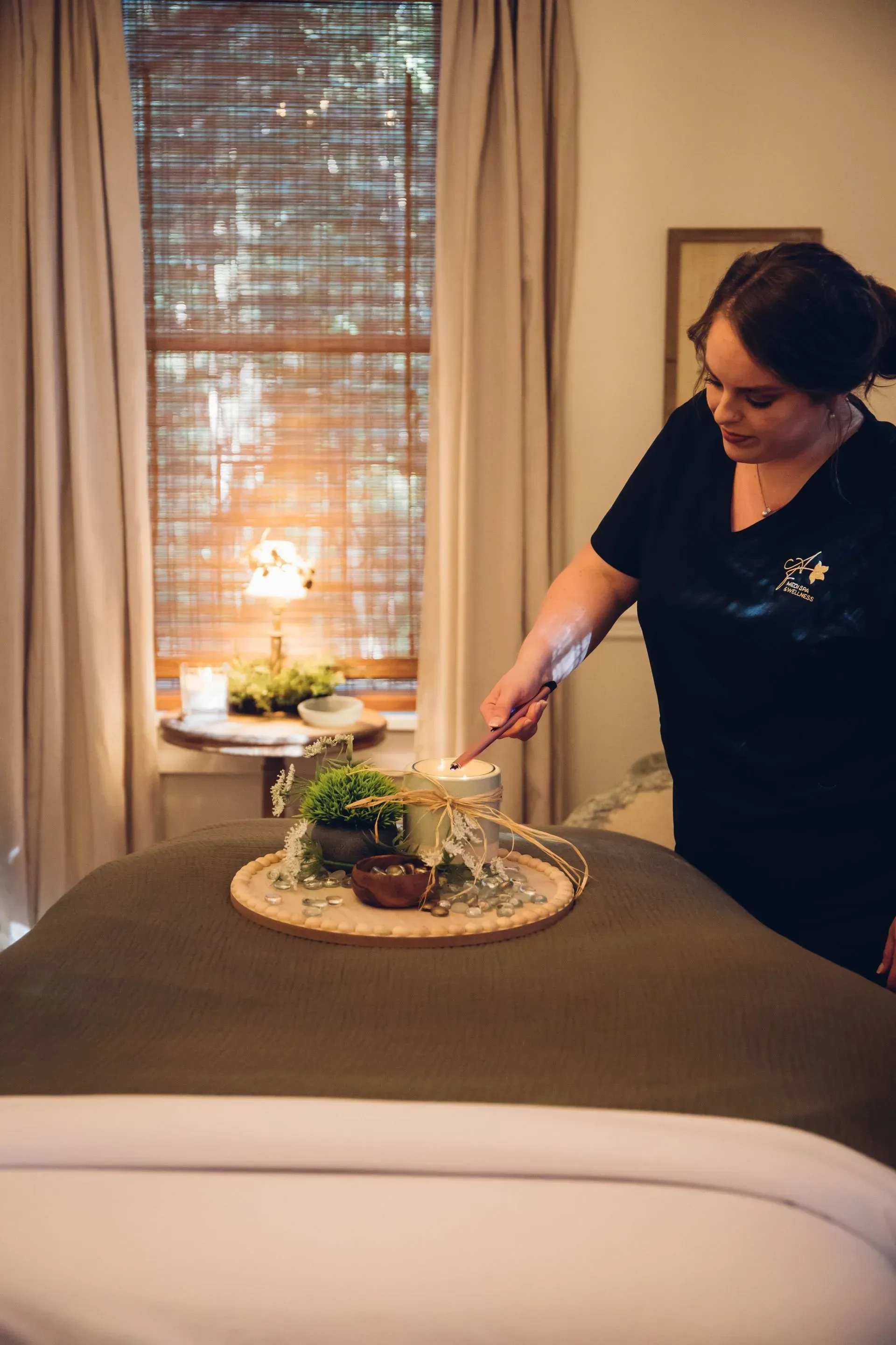 Woman preparing massage table with tray of plants and candle near window.