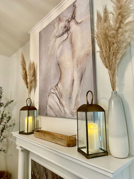 Mantel decorated with pampas grass, artwork of a figure, candles in lanterns, and a wooden box.