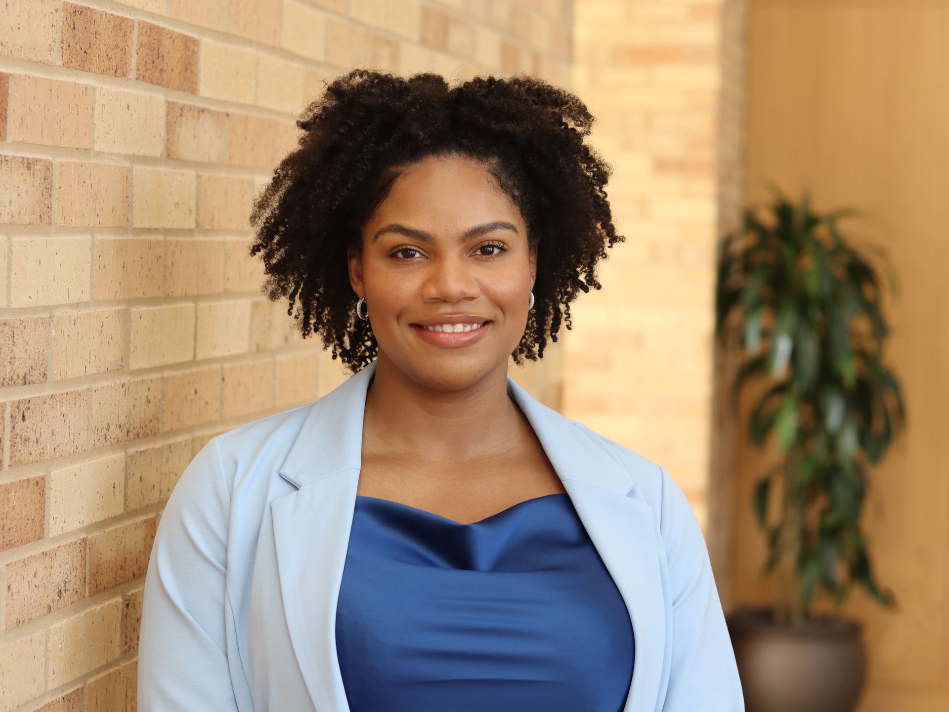 Smiling person in a light blue blazer standing indoors beside a brick wall and potted plant