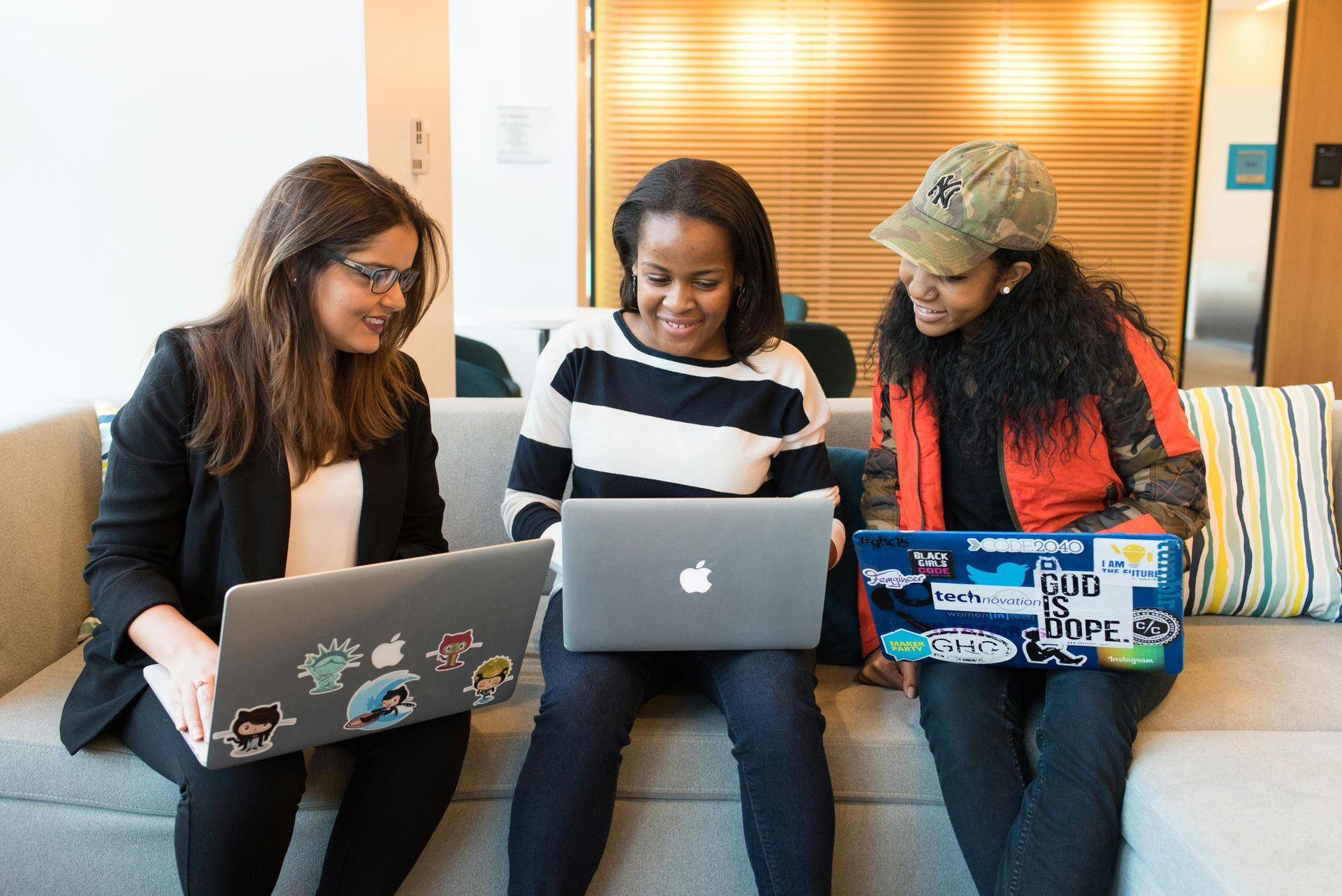 Three people sit on a couch using laptops in a bright lounge.