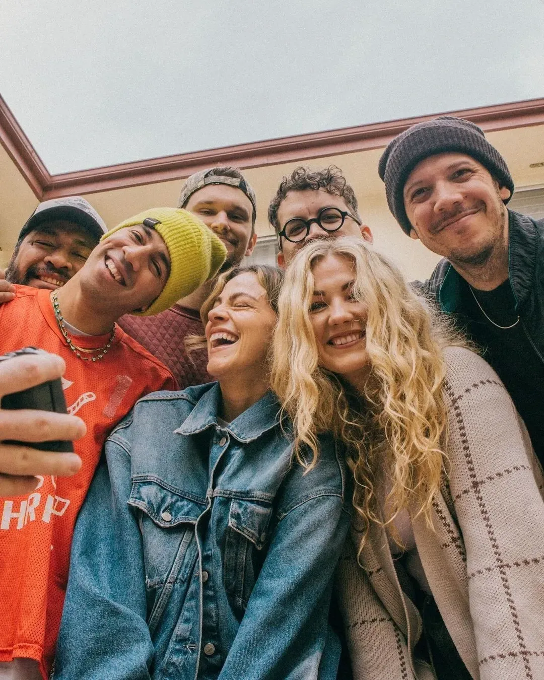 Six friends smiling and posing closely together outdoors, wearing casual sweaters and jackets.