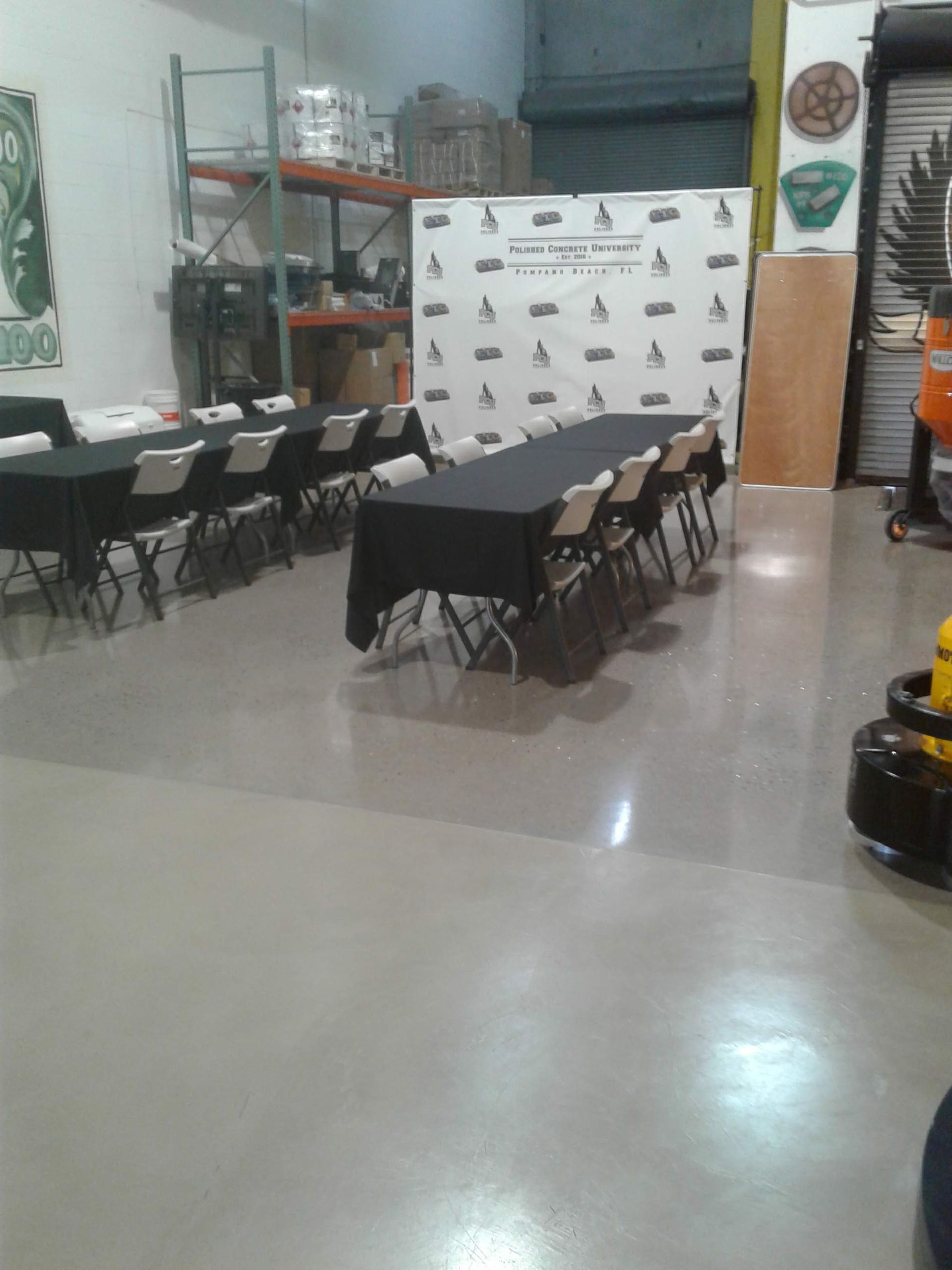 Two long tables with black tablecloths and white chairs set up for an event in a warehouse with a branded backdrop.
