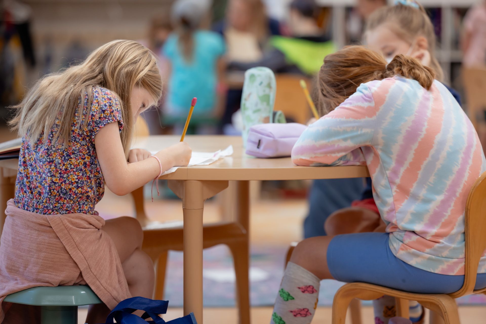 Two children lean over a table, drawing with pencils in a classroom or art activity area.