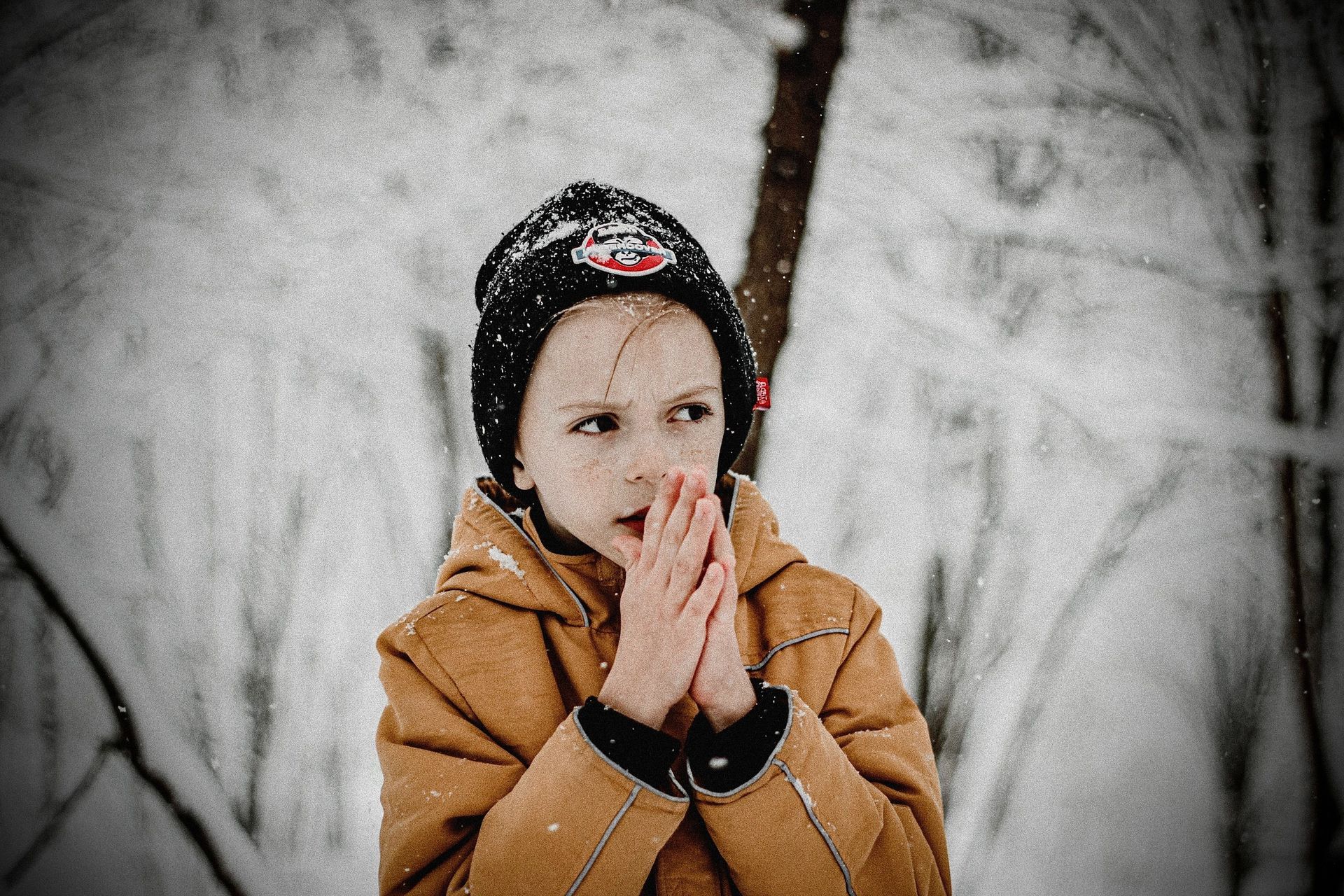 Child in a tan coat and black hat standing in a snowy forest, hands near face, looking worried.