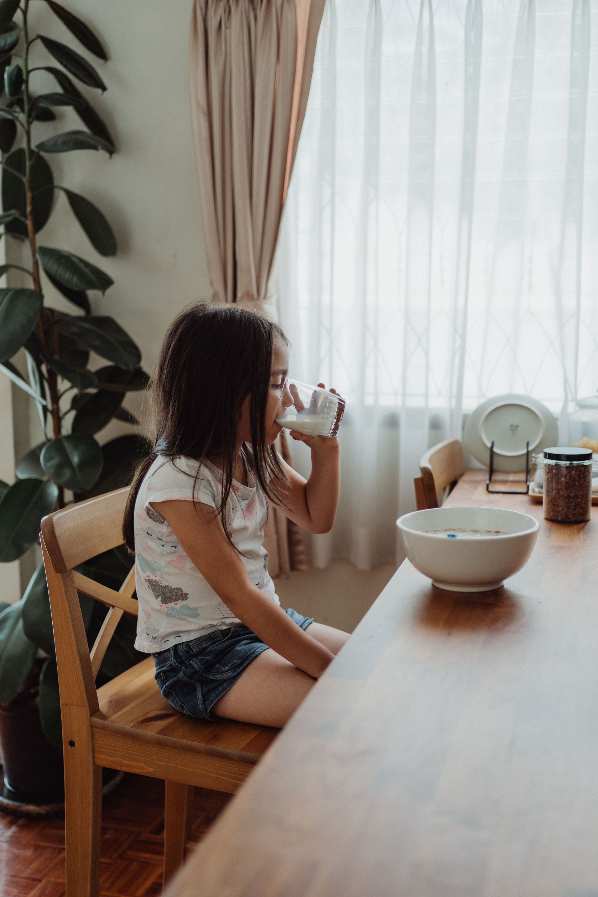 Child drinking at a wooden table beside a bowl in a bright room with sheer curtains
