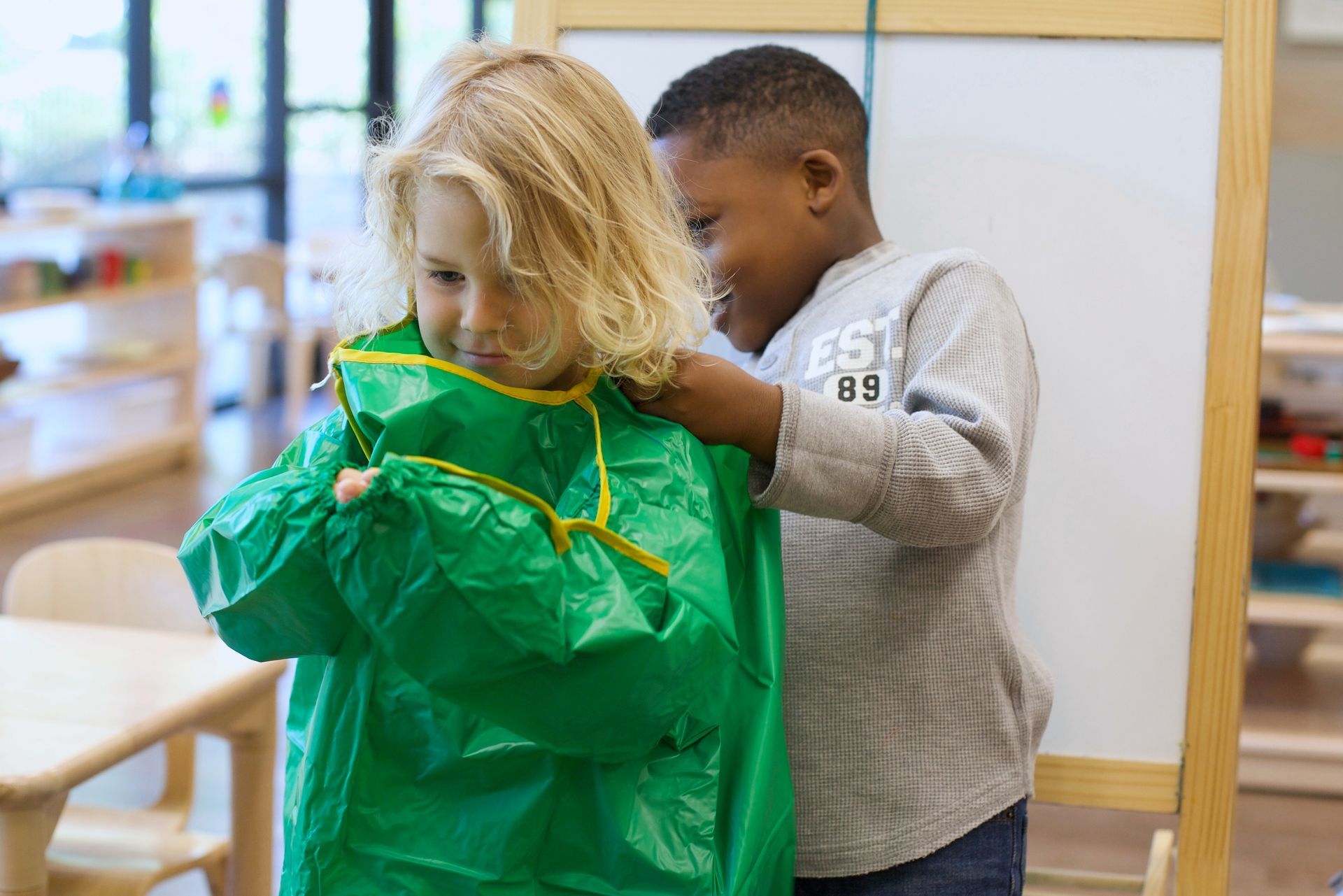 One child helping another child put on a green painting apron in a classroom