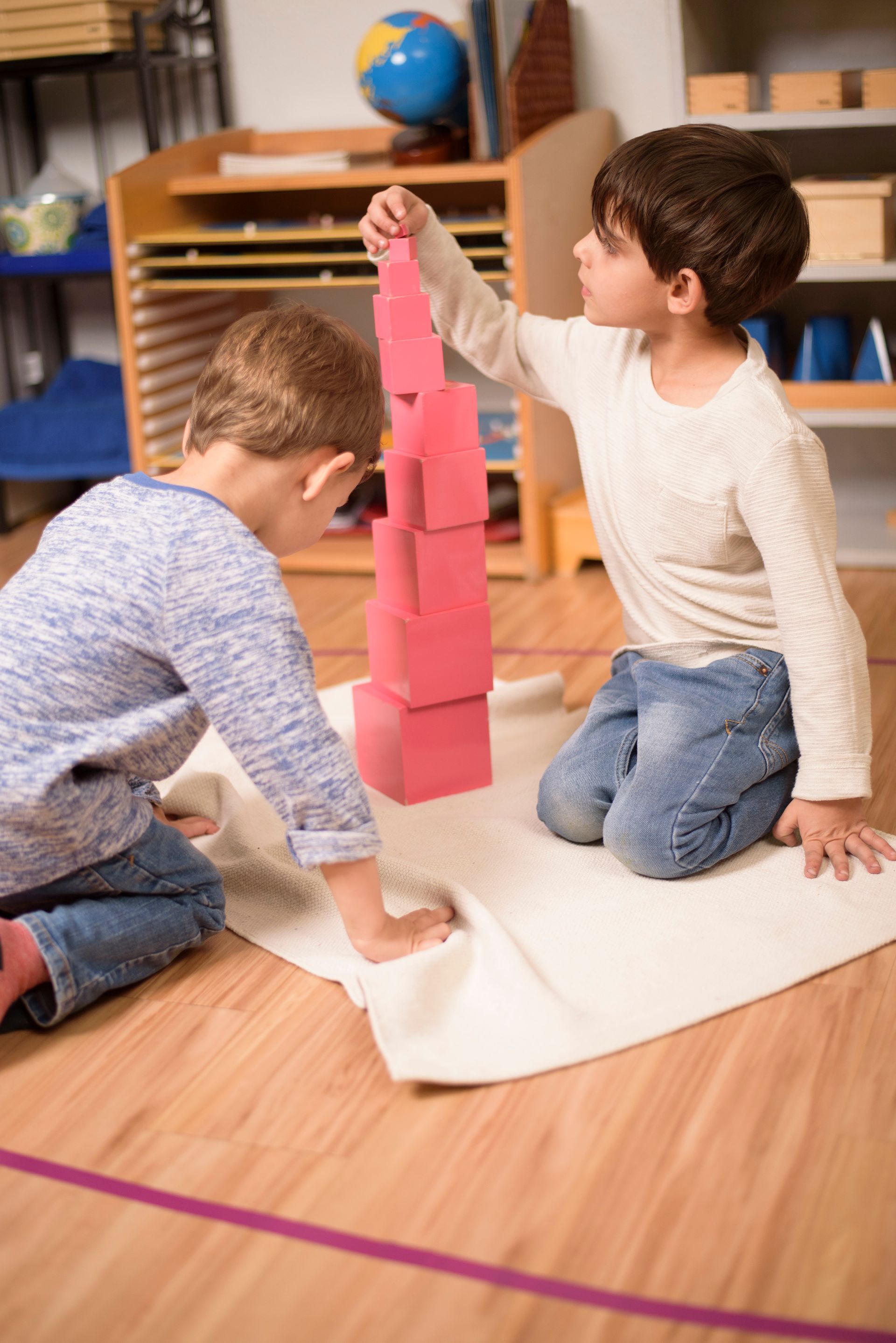 Two children kneel on a mat, stacking the Pink Tower in a classroom.