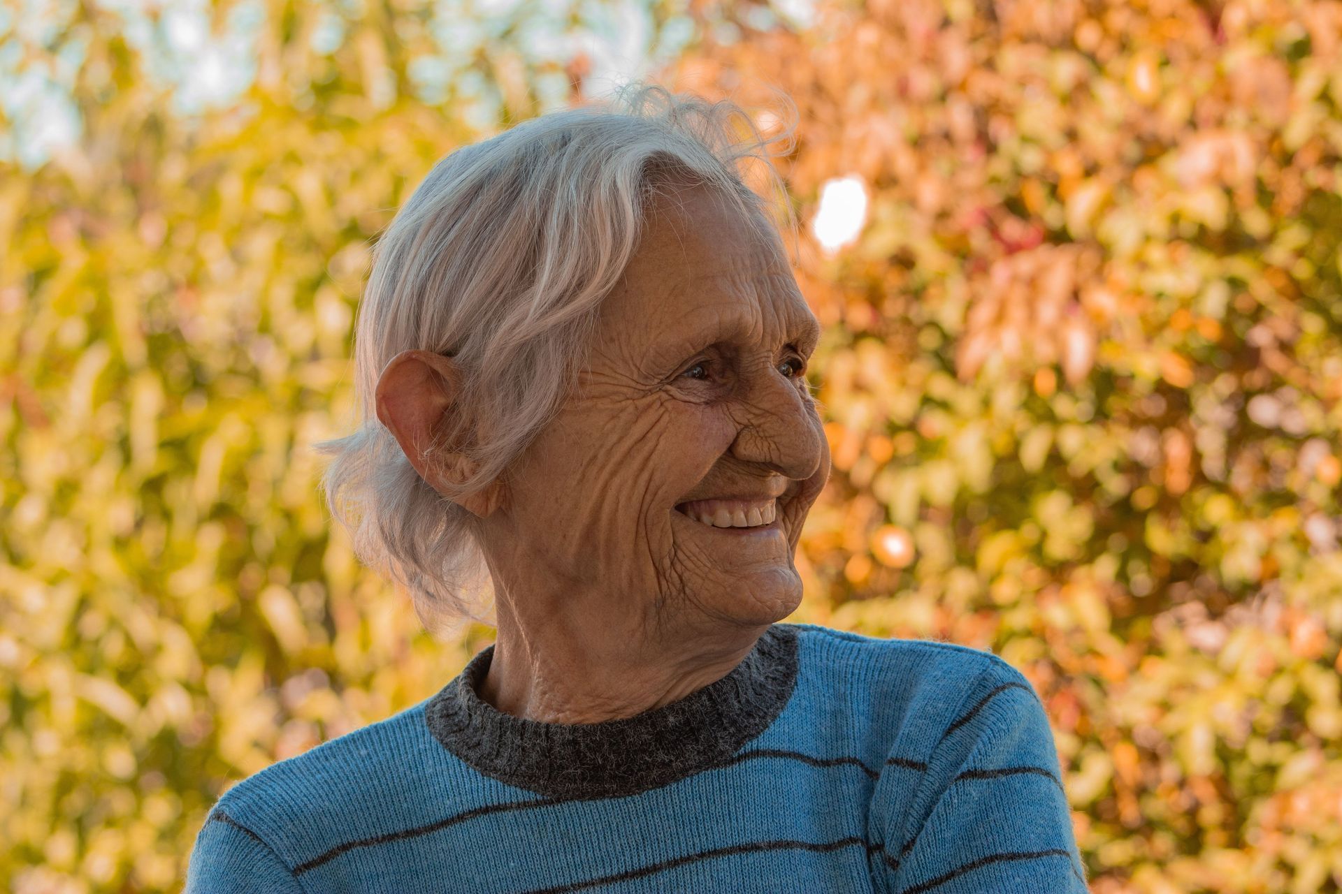 Smiling woman in a blue sweater outdoors with warm autumn foliage behind her