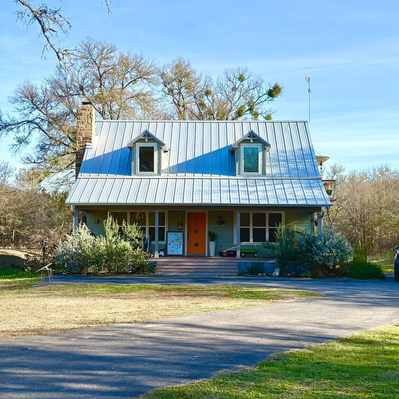 Rustic house with white metal roof, two dormer windows, and a front porch surrounded by trees and lawn.