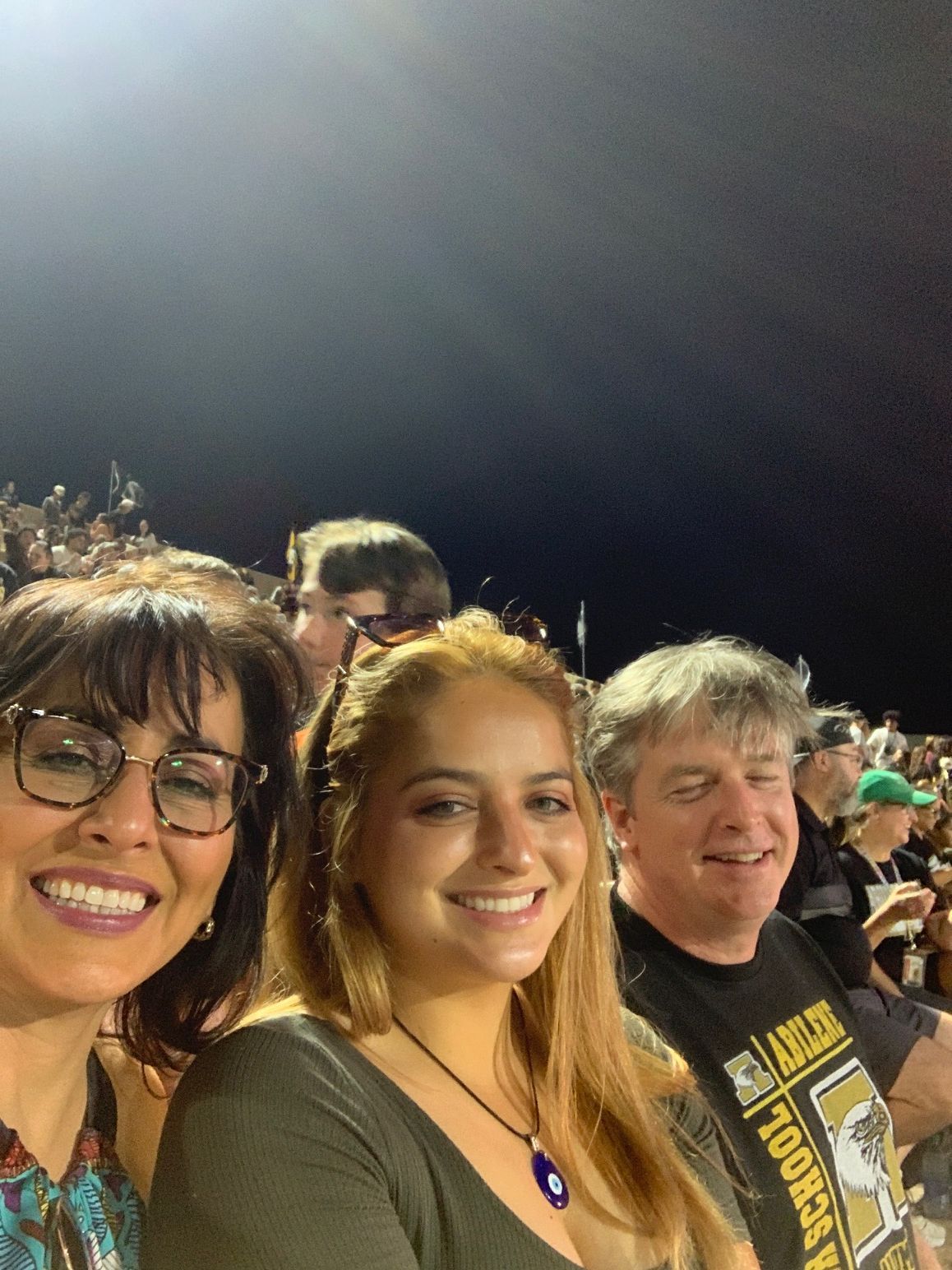 Three smiling people posing at a nighttime outdoor event, with stadium lights glowing overhead.