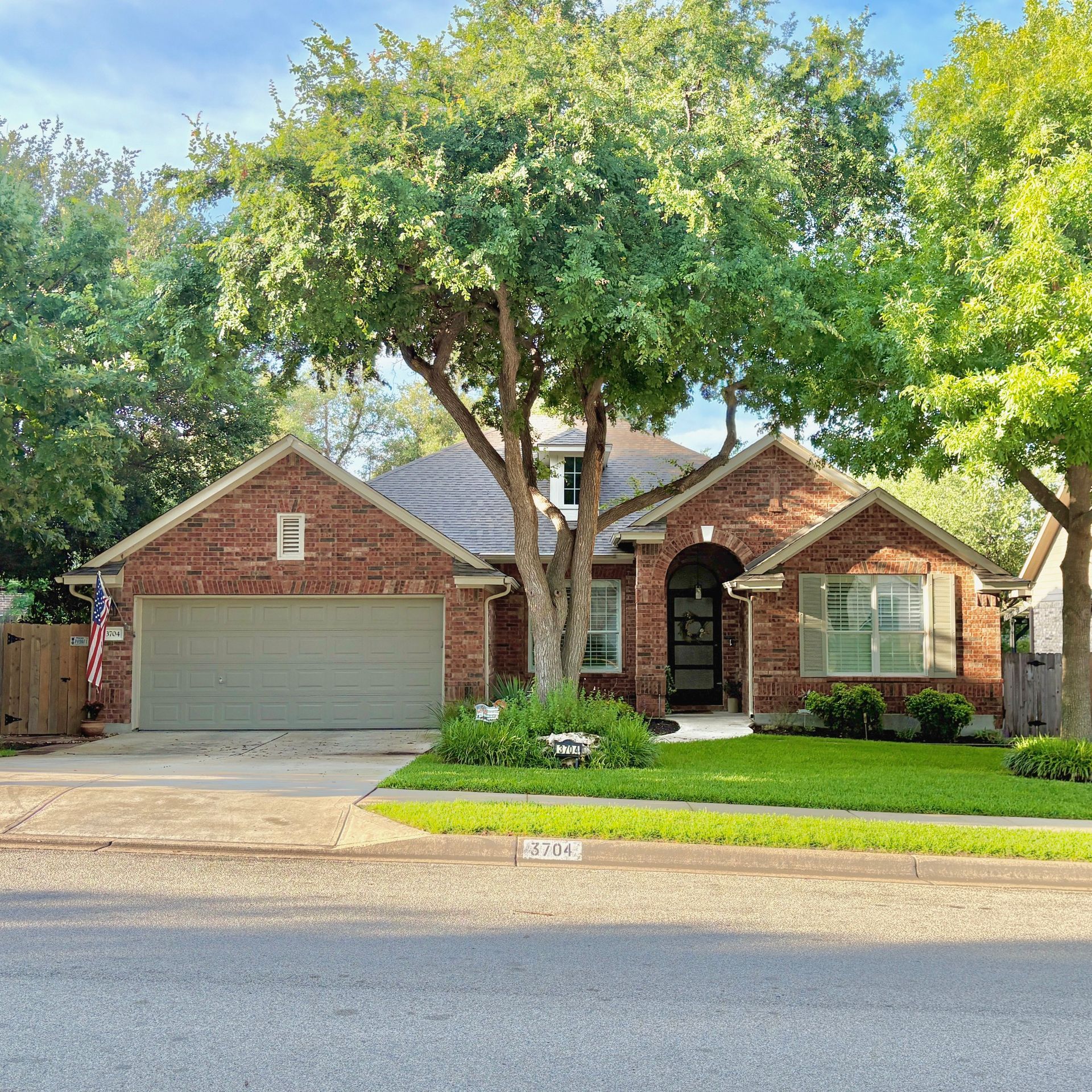 Brick suburban house with a two-car garage, large front tree, and neatly trimmed lawn