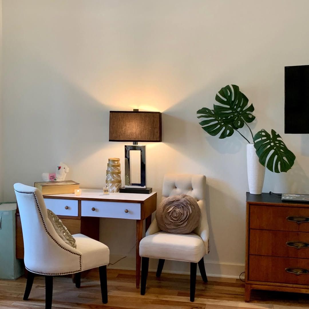 Cozy corner with a desk lamp, two cream chairs, wooden desk, cabinet, and large leafy plant.