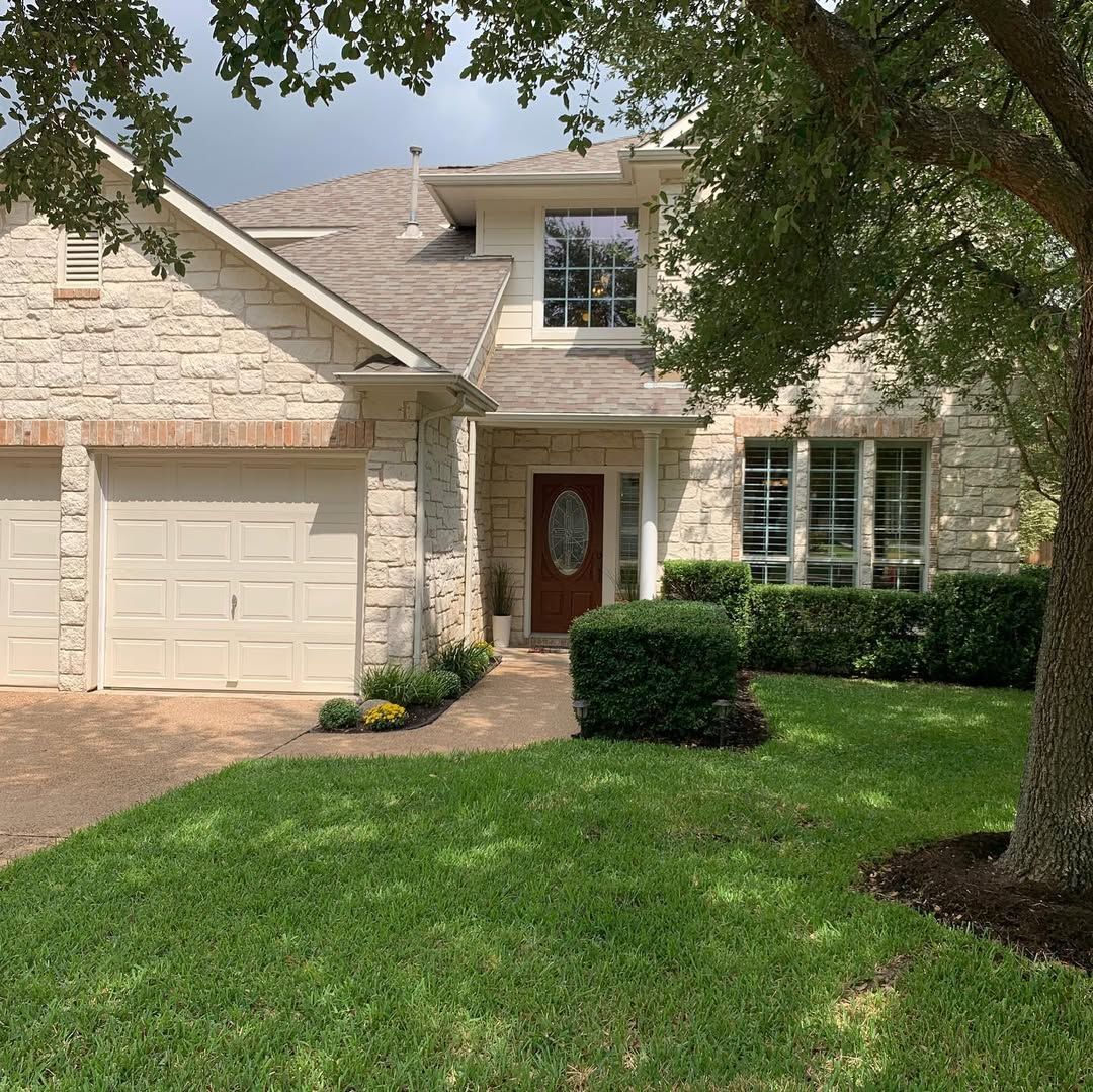 Suburban stone house with garage, red front door, and a green lawn shaded by trees