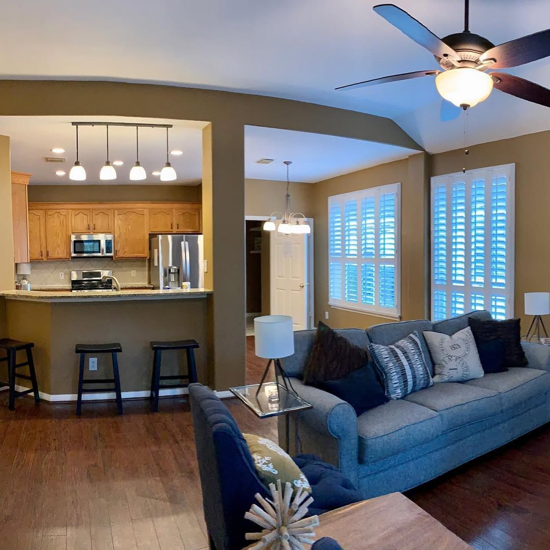 Open living room with blue sofa, kitchen bar stools, ceiling fan, and bright windows with shutters