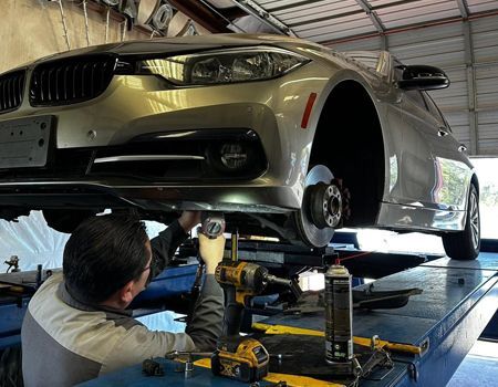 Mechanic working under a silver car on a lift in an auto shop, using tools to service the front wheel area