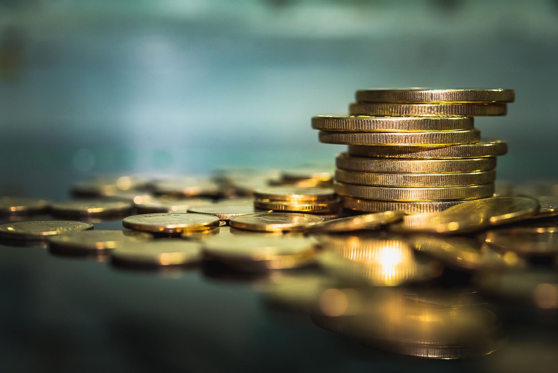 Stacked gold coins on a reflective surface with scattered coins in the foreground, blue blurred background