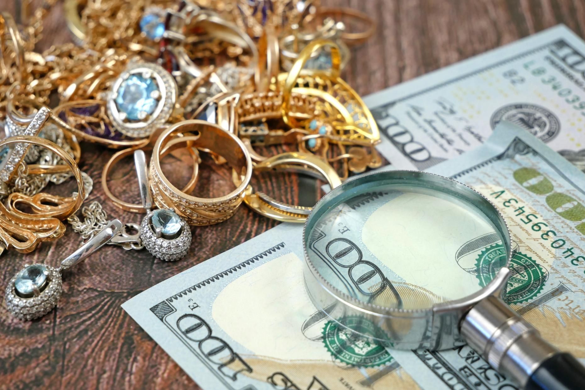 Gold jewelry and a magnifying glass over U.S. dollar bills, suggesting appraisal or inspection