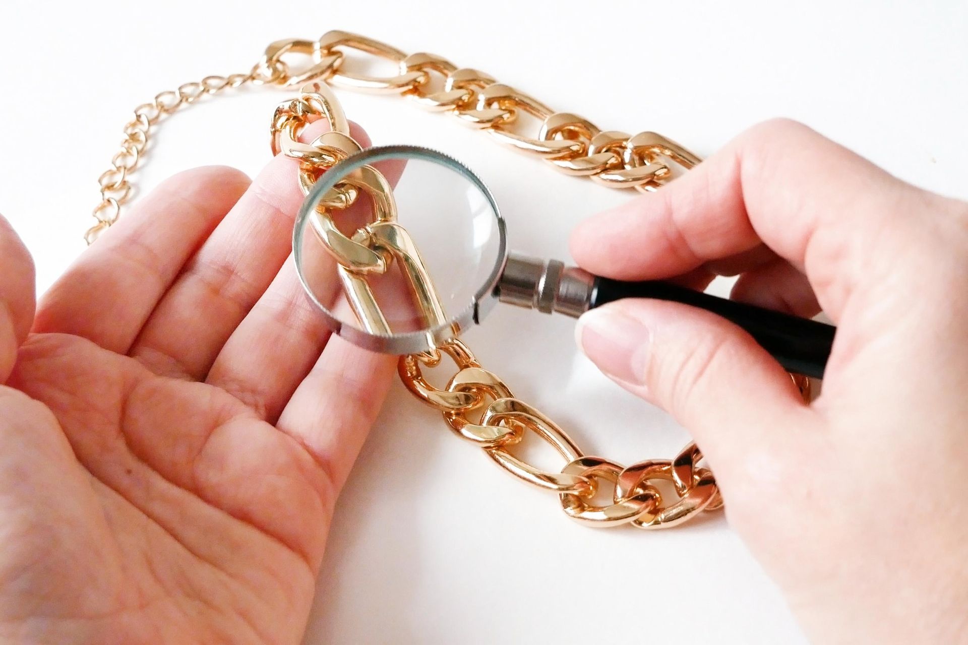 Hand holding a magnifying glass over a gold chain bracelet to inspect the jewelry