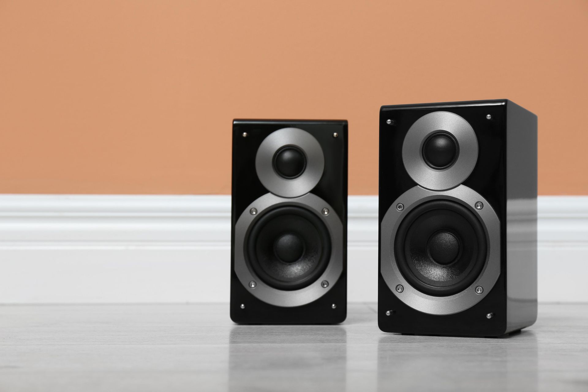 Two black bookshelf speakers on a light floor against a peach wall.