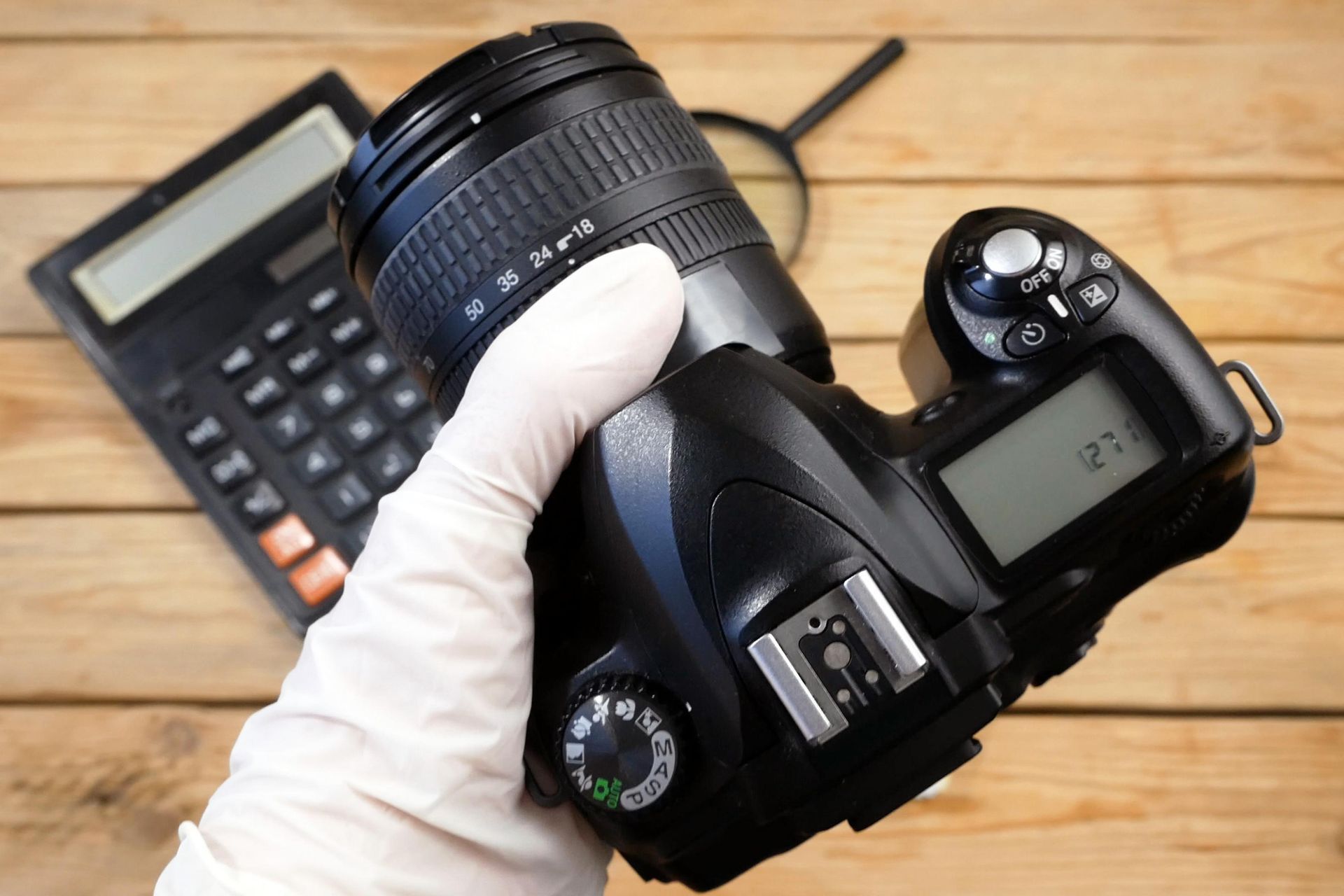 Gloved hand holding a DSLR camera over a calculator on a wooden desk