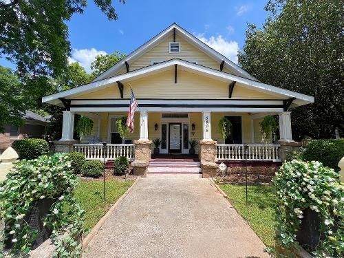 Yellow house with white trim, front porch, American flag, and brick walkway surrounded by greenery